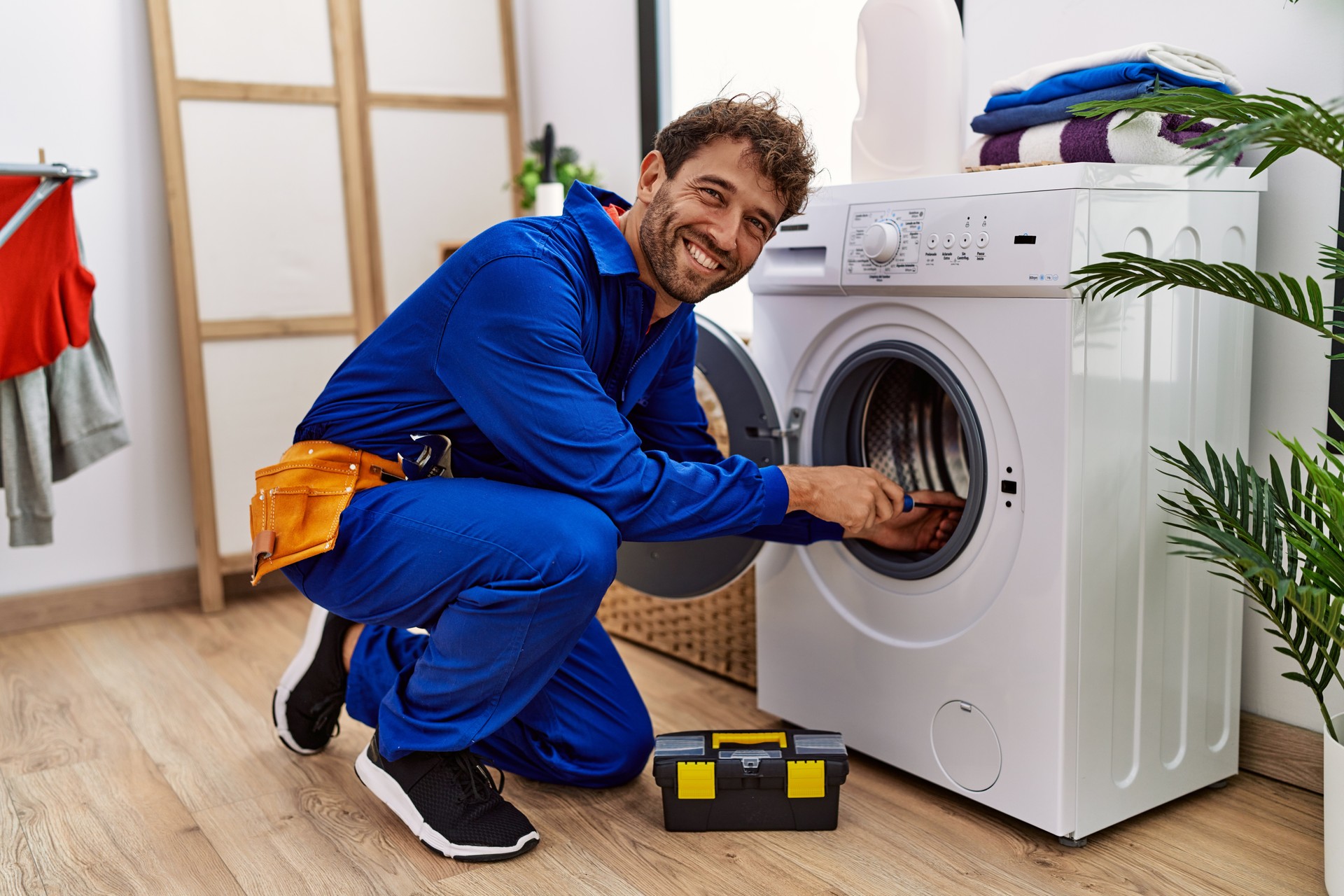 Young hispanic man wearing handyman uniform repairing washing machine at laundry room